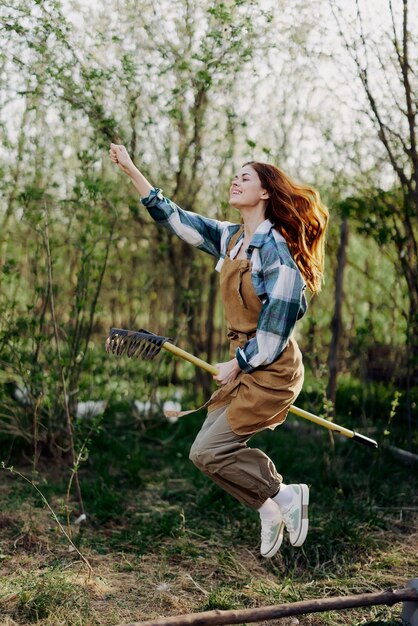 The woman hops amusingly with her rake like a witch on a broomstick in her green garden among the trees and smiles The concept of everyday lifestyle and joy in the moment