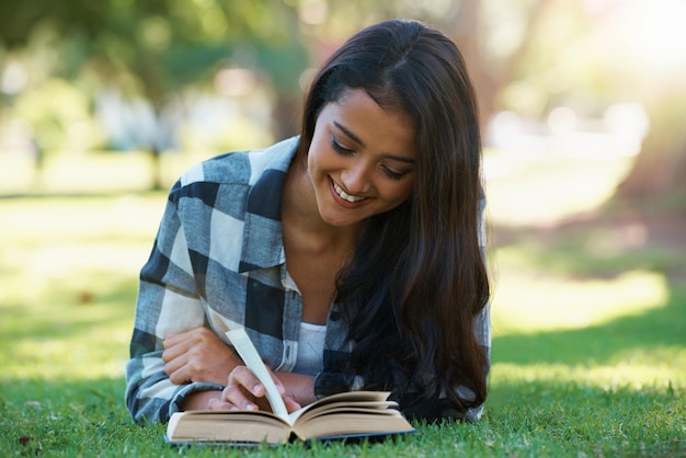 Woman information and reading book on grass literature and smiling for fiction or fantasy story Female person nature and relaxing on lawn for knowledge novel and student studying in garden