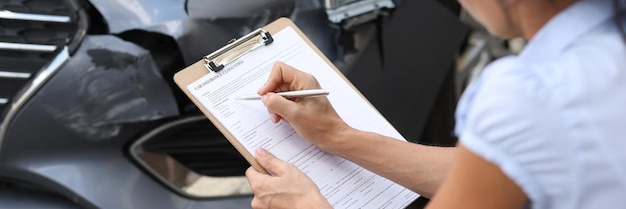 Woman insurance agent filling out paperwork near wrecked car closeup estimation of cost of