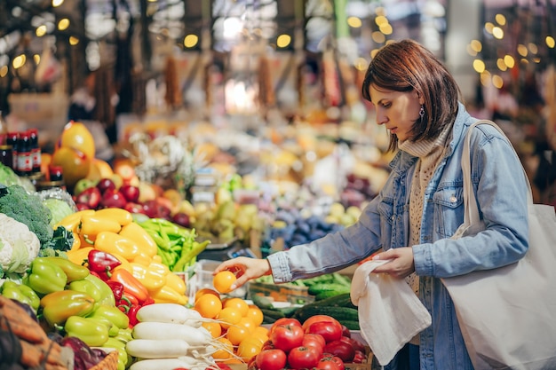 Woman is chooses fruits and vegetables at food market. Reusable eco bag for shopping. Sustainable lifestyle. Eco friendly concept.