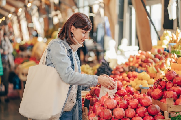 Woman is chooses fruits and vegetables at food market. Reusable eco bag for shopping. Sustainable lifestyle. Eco friendly concept.