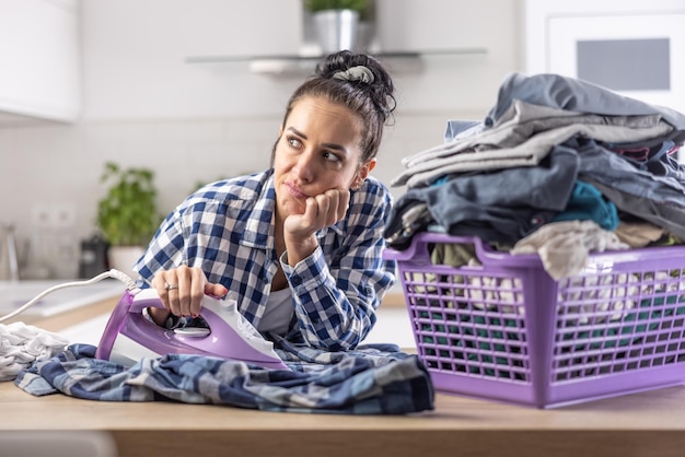 Photo woman is daydreaming while ironing next to an overfilled big basket full of clothes