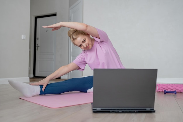 A woman is doing a home workout guided by a laptop stretching on a yoga mat in a cozy indoor