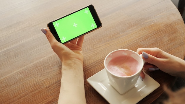 Woman is drinking a cup of coffee in cafe holding a mobile phone in another hand. Green screen, hands close-up.