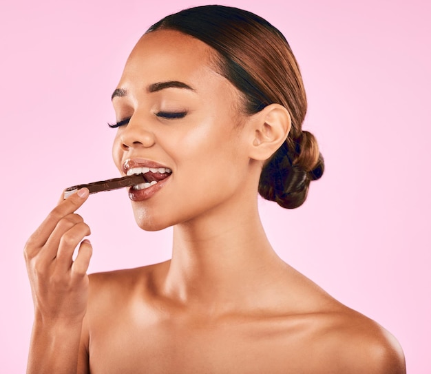 Woman is eating chocolate health and beauty with candy skincare and wellness on pink background Female model with sweets snack cacao dessert and glow with makeup and cosmetic shine in studio