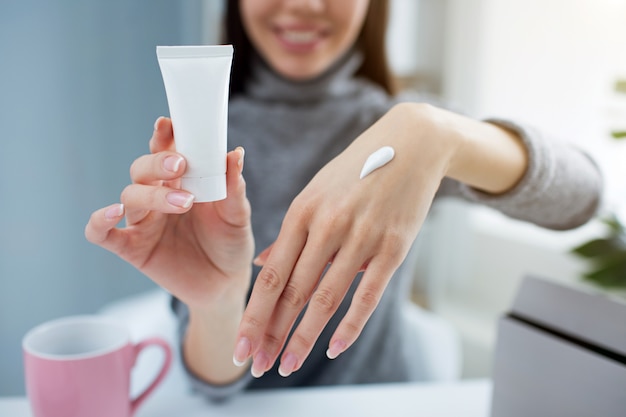 Woman is holding a small tube with hand cream in one hand and showing her other hand with some cream on it