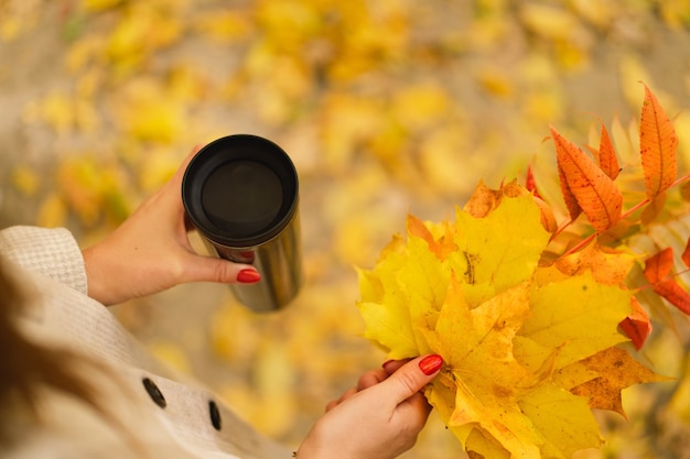 A woman is holding a thermo cup and a bouquet of autumn leaves autumn outdoor activity