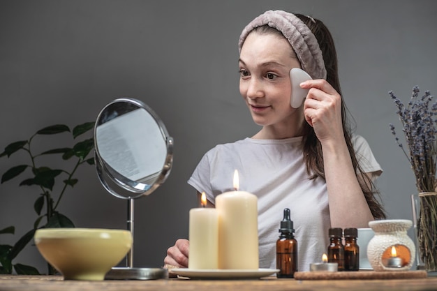 Woman is looking in the mirror and doing a massage with a gouache scraper Candles and an aroma lamp with essential oils on the table Concept of facial care aromatherapy morning and evening rituals