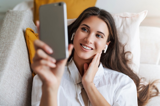 Woman is lying on couch, relaxing, using smartphone for text messages, exchanging photos, watching videos.