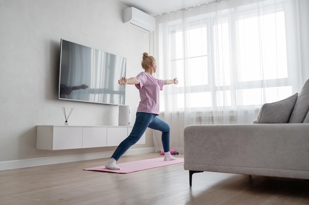 A woman is practicing yoga in a cozy living room on a pink mat doing a warrior pose with a serene