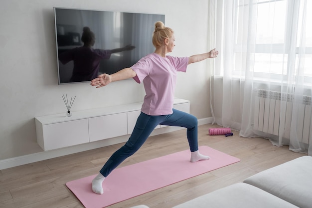 A woman is practicing yoga in a cozy living room on a pink mat doing a warrior pose with a serene