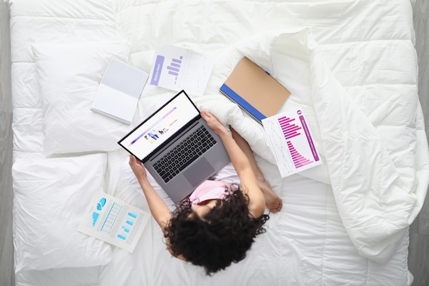 A woman is sitting on the bed with a laptop and documents.