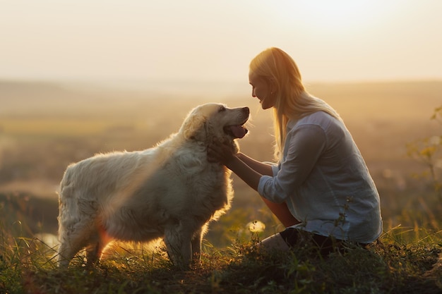 woman is sitting on the hill at sunset lovingly hugging her large breed dog