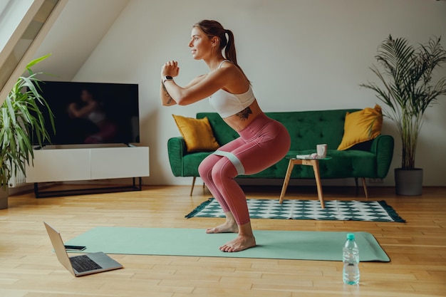 A woman is squatting on a yoga mat on hardwood flooring in a living room Home workout concept