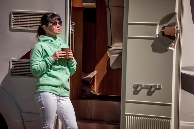 Woman is standing with a mug of coffee near the camper. Caravan car Vacation. Family vacation travel, holiday trip in motorhome RV