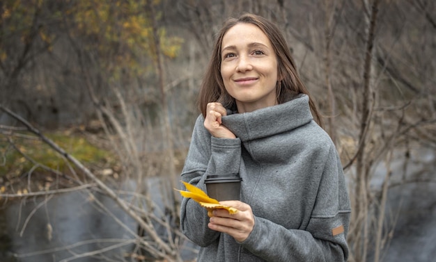 Woman is walking through the autumn forest along the river with a cup of coffee and enjoying the beautiful nature fresh air and calm