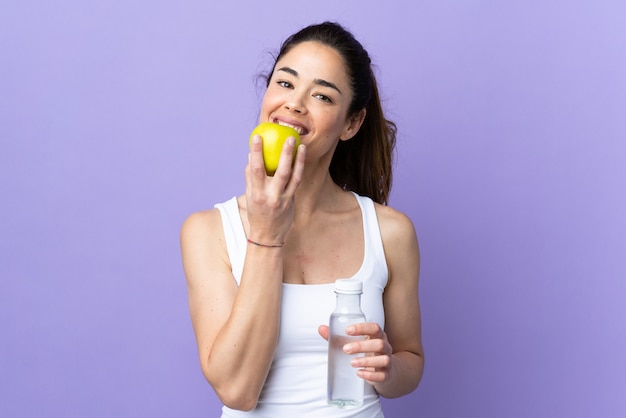 Woman over isolated purple with a bottle of water and eating an apple