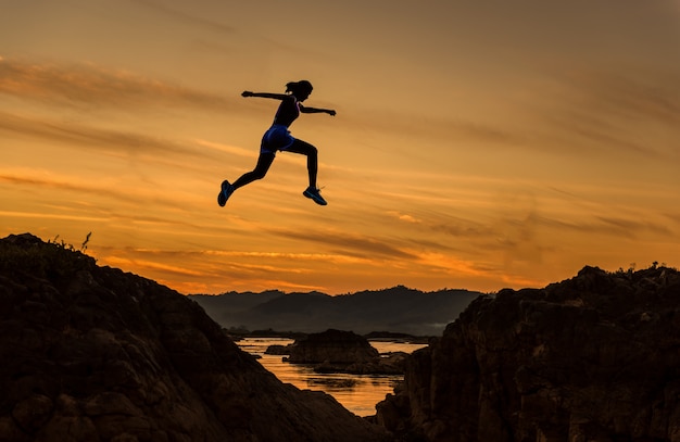 Woman jump through the gap between hill.woman jumping over cliff on sunset background