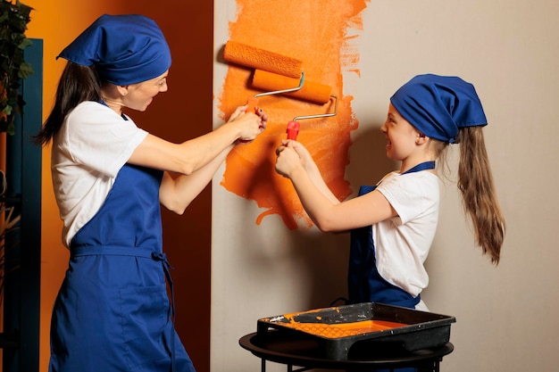 Woman and kid painting house walls with orange color paint, using renovating tools and paintbrush or roller to decorate apartment. Housework renovation with equipment, diy decoration.