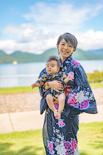 A woman in a kimono holds a baby in front of a lake.