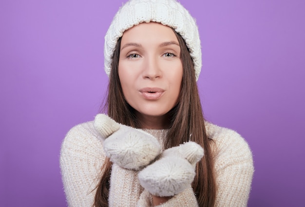 A woman in a knitted white hat and mittens blows a kiss.