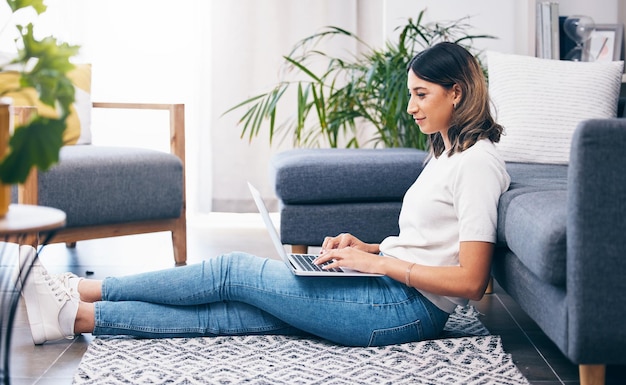 Woman laptop and typing sitting on floor in the living room by sofa in remote work or studying at home Female freelancer working on computer checking email research or browsing in lounge by couch