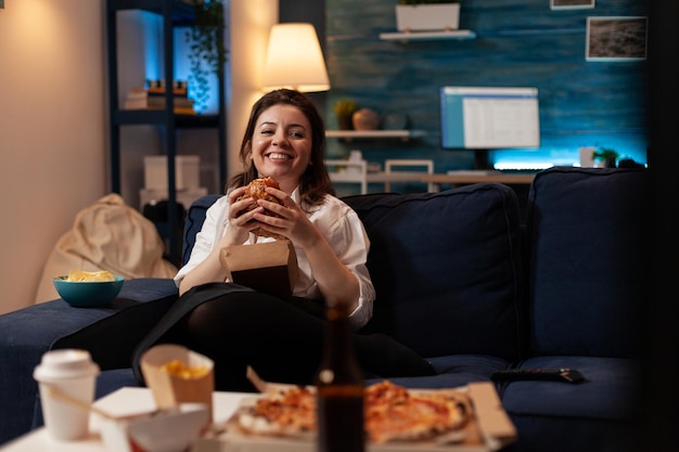 Woman laughing watching comedy television series while eating tasty takeout burger relaxing on couch in living room. Smiling person having takeaway fast food dinner in front of tv.