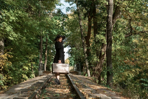 Woman leaves with a suitcase along the rails through the autumn forest Woman in black clothes Back view