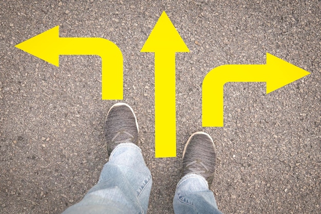 Photo woman legs in sneakers standing on asphalt road with three direction yellow arrows