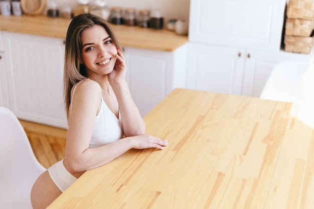 Woman in Lingerie Smiling Sitting at Wooden Table Blurred Kitchen in Background