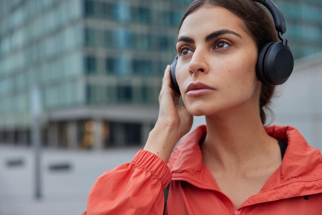 woman listens motivational music via headphones dressed in windbreaker thinks about something poses outdoor