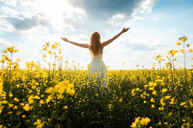 Woman in a long dress stands in a field with yellow flowers and raising her hands to the sun