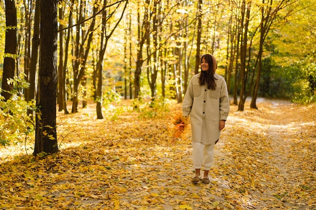 Woman in a long shirt with a bouquet of autumn leaves in her hands walks through the autumn forest