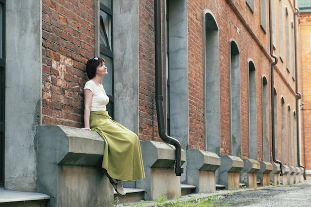 Woman in a long skirt sits on the basement cornice of an old building with large windows and a red brick facade