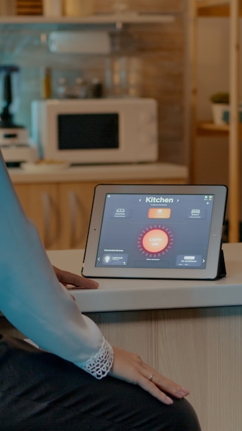 Woman looking at tablet in house with automation lighting system, sitting in kitchen switching on bulbs using voice command