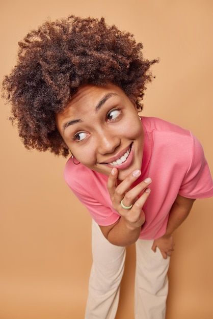 woman looks aside with cheerful smile keeps hand on chin being in good mood dressed casually feels very happy poses against brown expresses sincere emotions