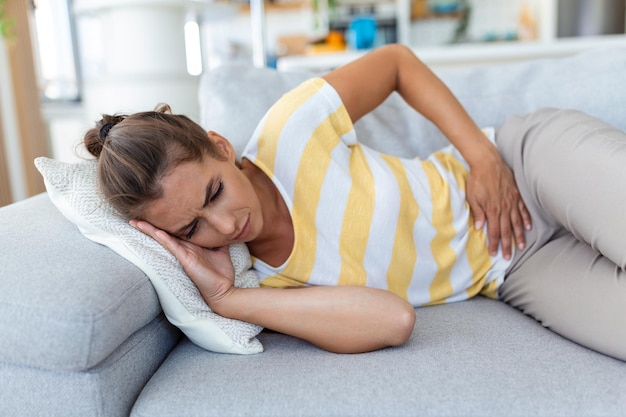 Woman lying on sofa looking sick in the living room Beautiful young woman lying on bed and holding hands on her stomach Woman having painful stomachache on bed Menstrual period