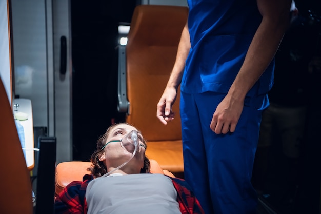 A woman lying on a stretcher with an oxigen mask on and looking at the paramedic in a blue uniform.