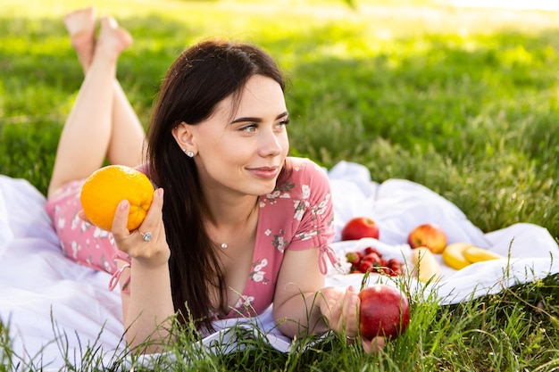 Woman lying on white bedsheet smiling and hold apple and orange in hand Lady have a rest