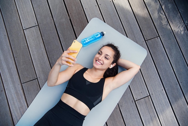 Woman lying on yoga mat after exercising relaxing with smartphone and listening to music workout