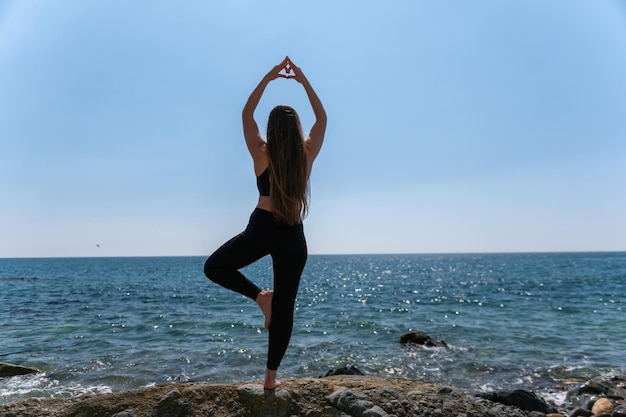 Woman makes heart with hands on beach Young woman with long hair fitness instructor stretching before pilates on a yoga mat near the sea on a sunny day female fitness yoga routine concept