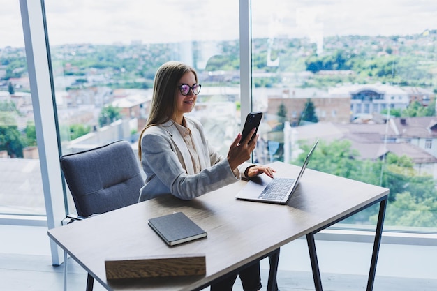 A woman manager in glasses sits at a laptop in an office with a stunning view of the city and conducts a business conversation on the phone Business lady working at the wooden table in the office