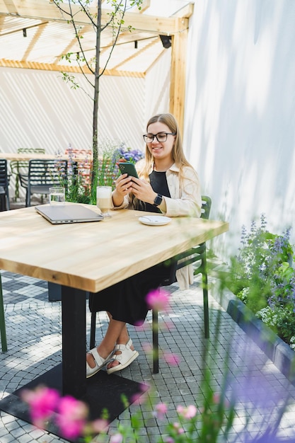 A woman manager sits on an outdoor terrace in a cafe and works online with coffee Female freelancer works remotely online while sitting in a summer cafe Remote work