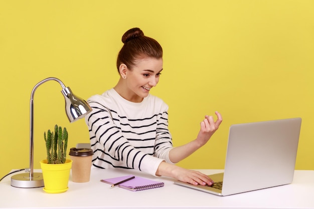 Woman manager sitting at workplace and showing to laptop screen and making beckoning gesture