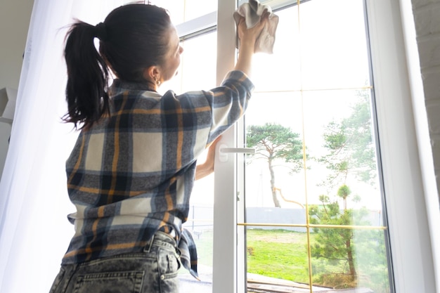 Woman manually washes the window of the house with a rag with spray cleaner and mop inside the interior with white curtains Restoring order and cleanliness in the spring cleaning servise