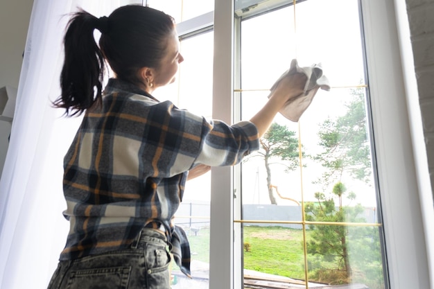 Woman manually washes the window of the house with a rag with spray cleaner and mop inside the interior with white curtains Restoring order and cleanliness in the spring cleaning servise