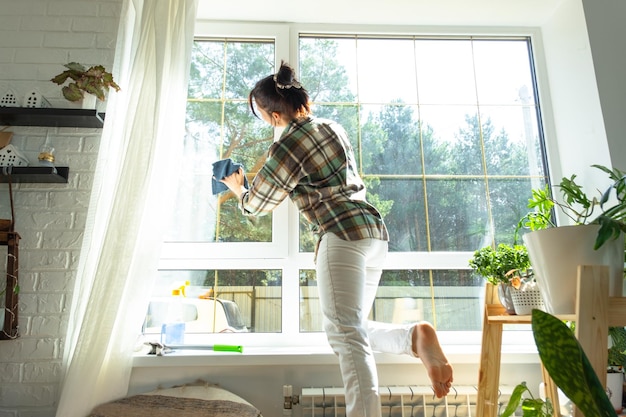 Woman manually washes the window of the house with a rag with spray cleaner and mop inside the interior with white curtains Restoring order and cleanliness in the spring cleaning servise