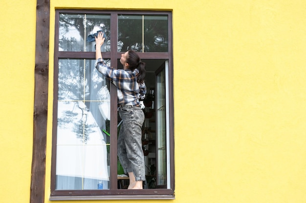 A woman manually washes the window of the house with a rag with a spray cleaner and a mop outside Safety at height restoring order and cleanliness in the spring cleaning service