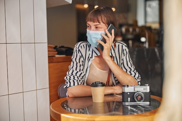 Woman in mask who is looking away while talking on smartphone and sitting in cafe