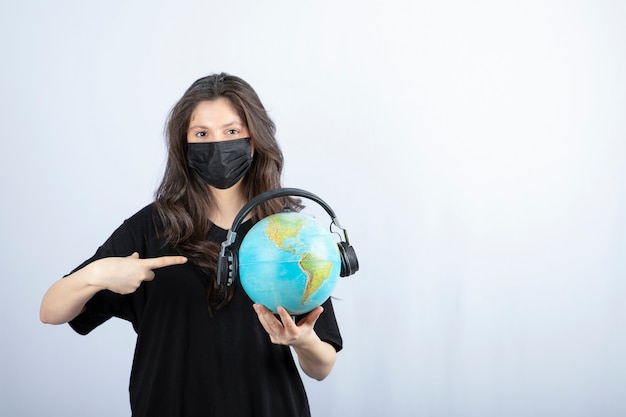 Woman in medical mask holding and pointing to globe on white table. 